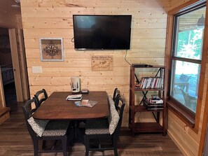 Interior em madeira com mesa de jantar, cadeiras, estante, televisão na parede e janela com vista.