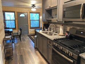 A kitchen with a stove, sink, and cabinets, a dining area with chairs and a table, and a view through the windows.