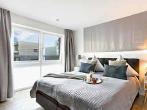 A modern bedroom with a large bed, a bedside table, a window with curtains, and a view of buildings outside. - NEW - Sky 65 Plus (Kappeln)
