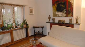 A living room with a white sofa, a radiator, a table with decorative items, a painting on the wall, and a window with curtains.