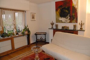 A living room with a white sofa, a radiator, a table with decorative items, a painting on the wall, and a window with curtains. - Holiday Residence in North Vosges (Bitche)