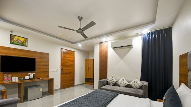 A hotel room with a bed, a desk with a TV, a wardrobe, a sofa with floral cushions, and a ceiling fan. - Hotel Rise Free Airport Pickup  (New Delhi)