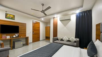 Quarto de hotel com cama, escrivaninha com televisão, guarda-roupa, sofá com almofadas florais e ventoinha no teto.