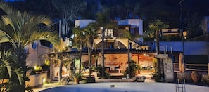 Un edificio de varios pisos con piscina, palmeras e iluminación exterior.