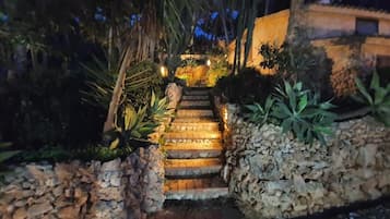 Eine Steintreppe führt zu einem Gebäude, umgeben von tropischen Pflanzen und beleuchtet mit warmem Licht.