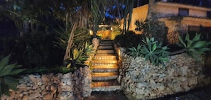 Una escalera de piedra conduce a un edificio, rodeado de plantas tropicales e iluminado con una luz cálida.