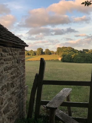 A rustic stone building with a wooden fence and bench, overlooking a grassy field and trees under a partly cloudy sky.