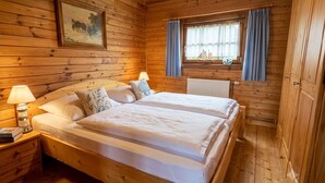 Habitación de cabaña de madera con cama doble, mesita de noche con lámpara, ventana con cortinas e imagen en el marco en la pared.