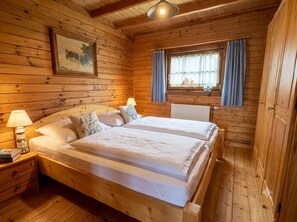 Habitación de cabaña de madera con cama doble, mesita de noche con lámpara, ventana con cortinas e imagen en el marco en la pared.