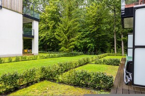 Un bâtiment moderne doté d’une façade en verre, d’un jardin bien entretenu avec des haies et des arbres, et d’une terrasse en bois.