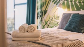 A neatly made bed with rolled towels, a patterned bedspread, and a large window with a view of greenery.