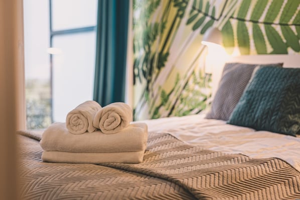 A neatly made bed with rolled towels, a patterned bedspread, and a large window with a view of greenery.