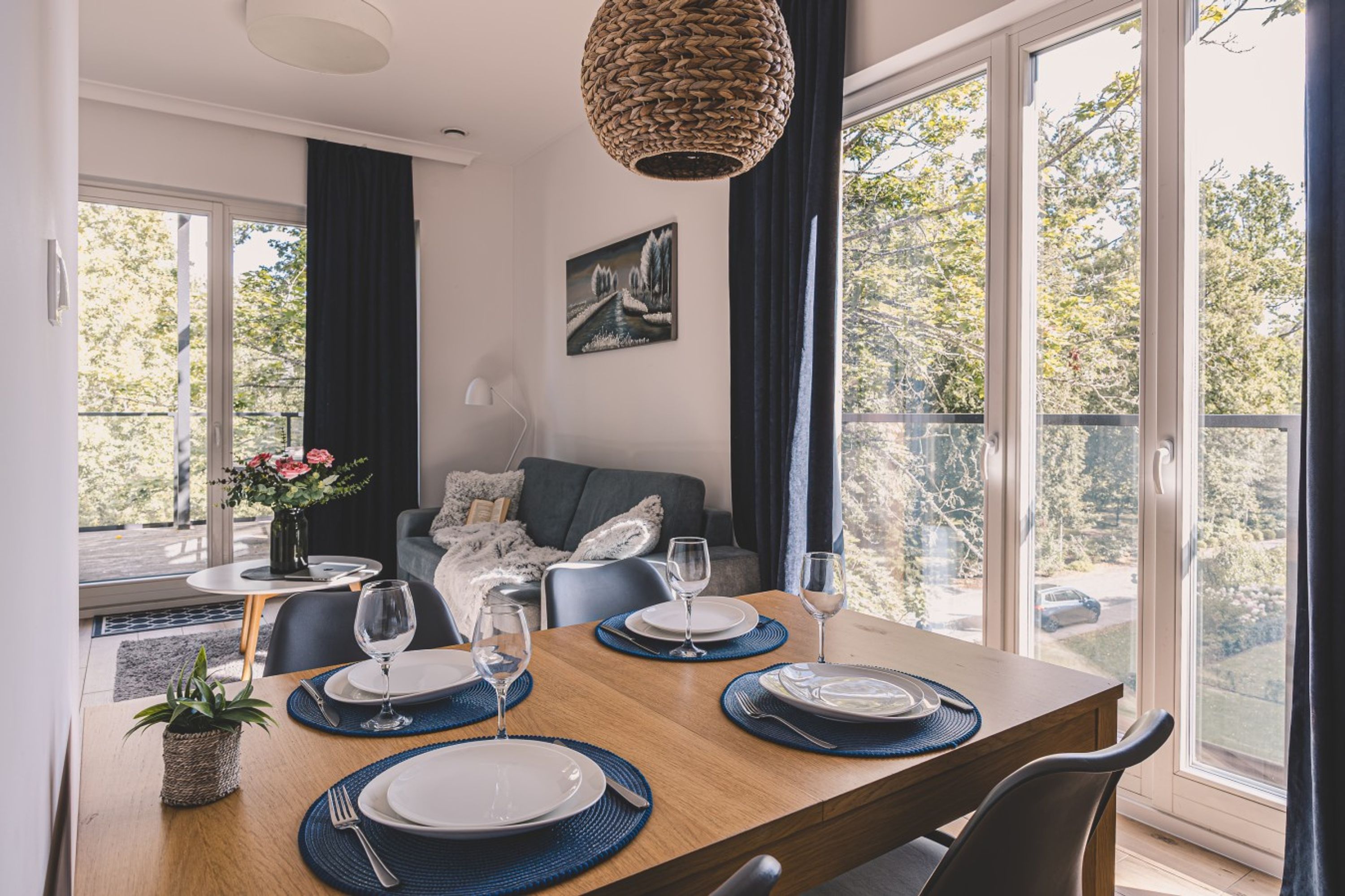 Un comedor con una mesa de madera dispuesta para cuatro personas, un sofá y un cuadro en la pared.