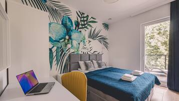 A modern bedroom with a bed, a desk with a laptop, and a large window.