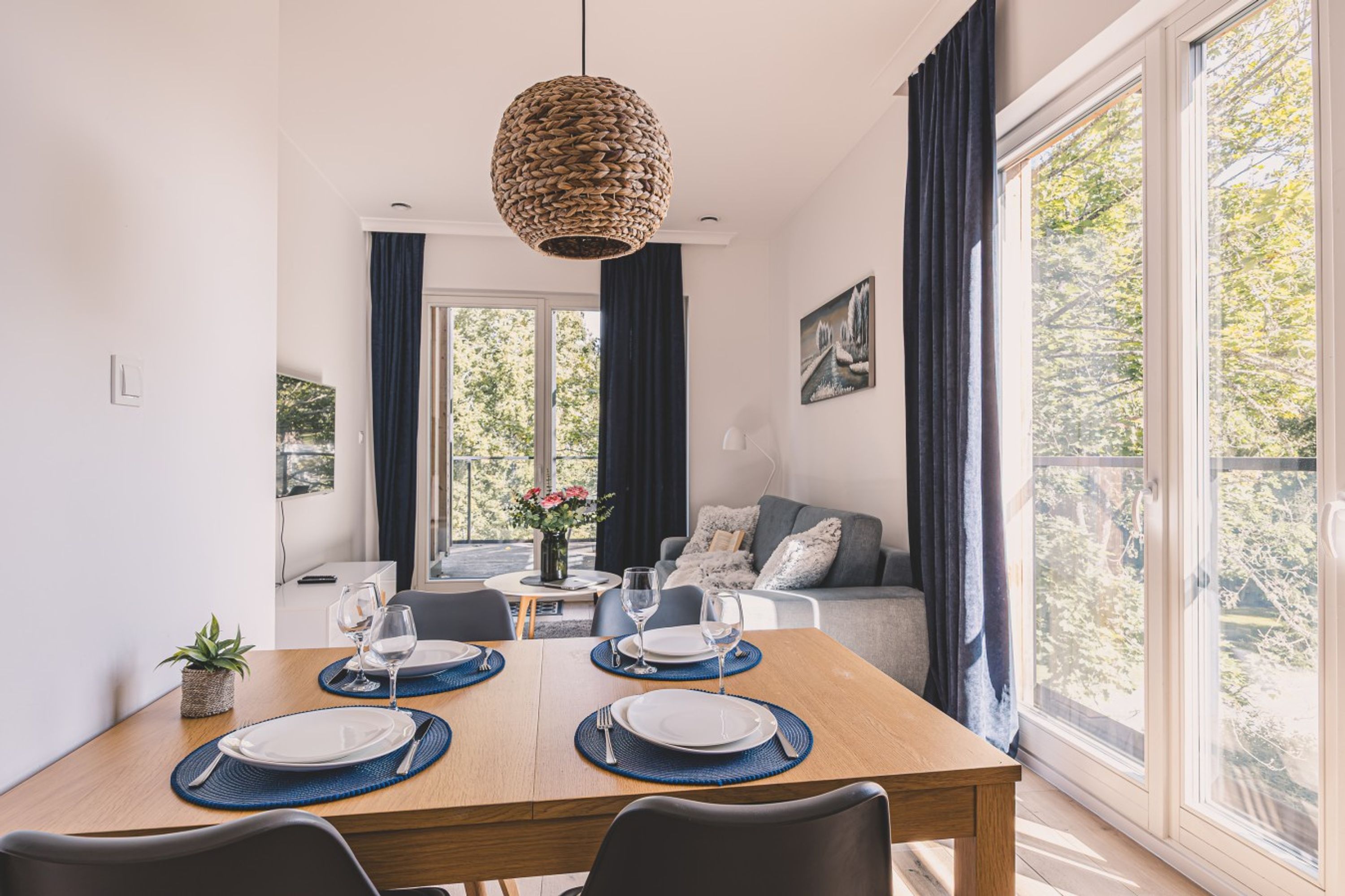 Un comedor con una mesa de madera dispuesta para cuatro personas, una lámpara colgante de fibras naturales y vistas al exterior a través de amplios ventanales.