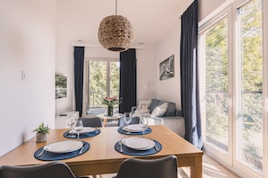 Un coin repas avec une table en bois dressée pour quatre personnes, un luminaire suspendu en fibres tressées et une vue sur l’extérieur grâce à de grandes fenêtres.