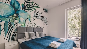 Un dormitorio con cama, mesita de noche, un ventanal con vistas a árboles y una pared con un mural de hojas tropicales.
