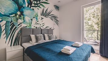 A bedroom with a bed, a nightstand, a large window with a view of trees, and a wall with a tropical leaf mural.