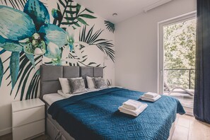 Une chambre à coucher comprenant un lit, une table de chevet, une grande fenêtre donnant sur la forêt et un mur orné d’une fresque représentant des feuilles tropicales.