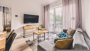 A modern living room with a flat-screen TV, a sofa, a dining table, and a sliding glass door leading to a balcony. - Bisonte Apartament 2 Sypialnie Du y Taras (Hajnowka)