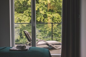 A room with a bed, a window with a view of greenery, and a chair outside.