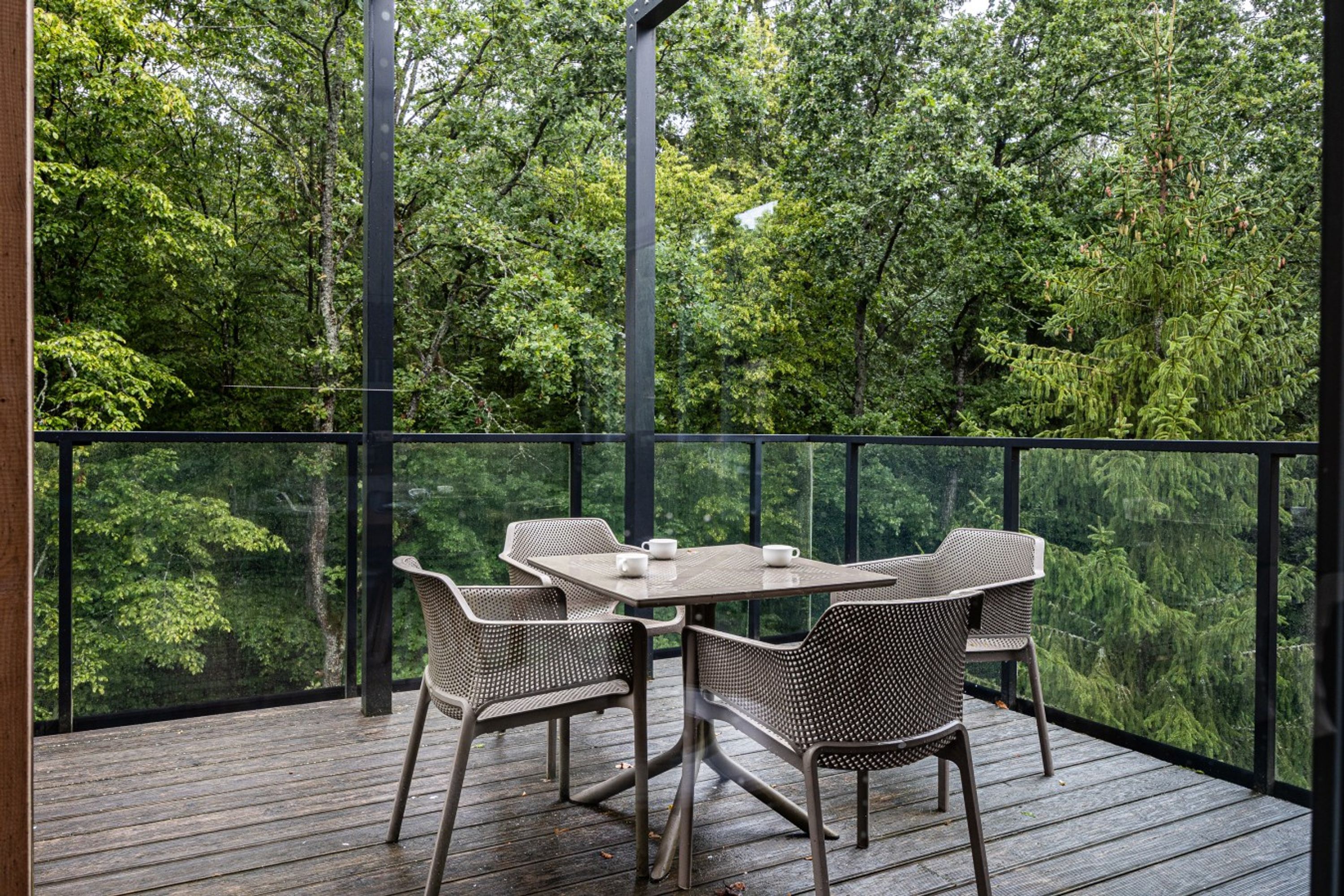 Una terraza exterior con barandilla de vidrio, una mesa y cuatro sillas.