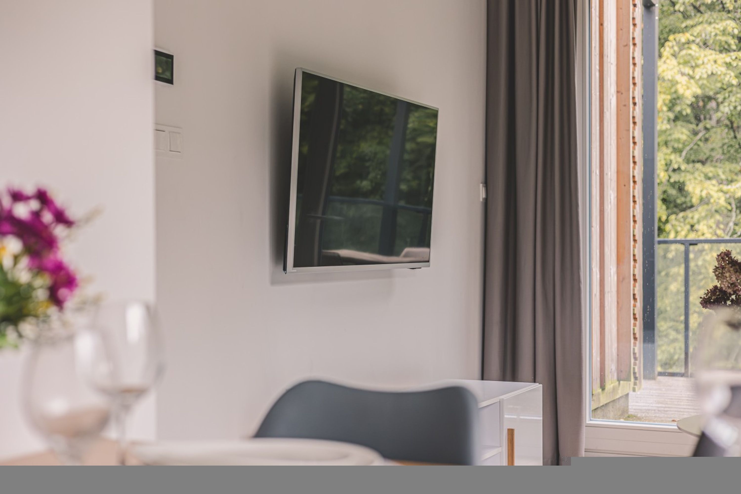 A modern room with a flat-screen TV mounted on the wall, a dining table with chairs, and a view of greenery through the window.