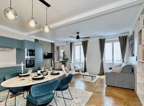 A modern kitchen with built-in appliances, a dining area with a round table and chairs, a grey sofa, and a ceiling fan. - Family flat for 6 steps from Champs Elysees (Paris)