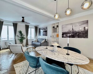 Un salon moderne avec une table ronde en marbre, des chaises bleues, un canapé gris et un tableau représentant un paysage urbain.