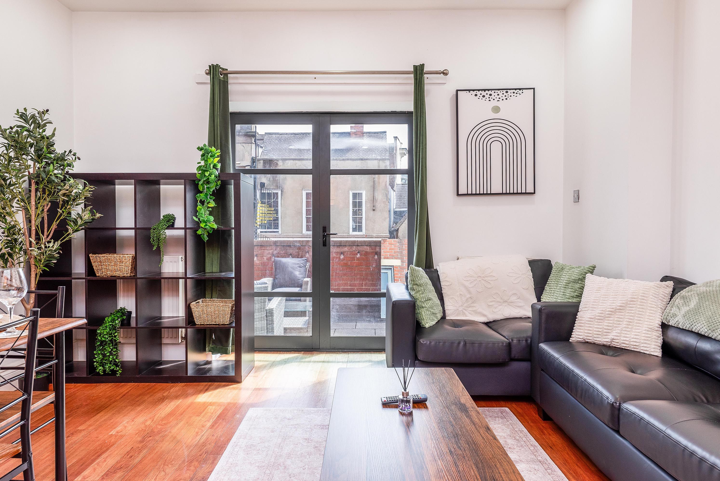 A modern living room with a black leather sofa, a glass coffee table, and a bookshelf with green plants.
