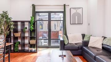 A modern living room with a black leather sofa, a glass coffee table, and a bookshelf with green plants.