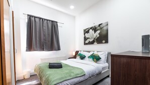 Ein Schlafzimmer mit einem Bett, einem Heizkörper, einem Fenster mit Vorhängen, einem Blumenbild und einem Schreibtisch.