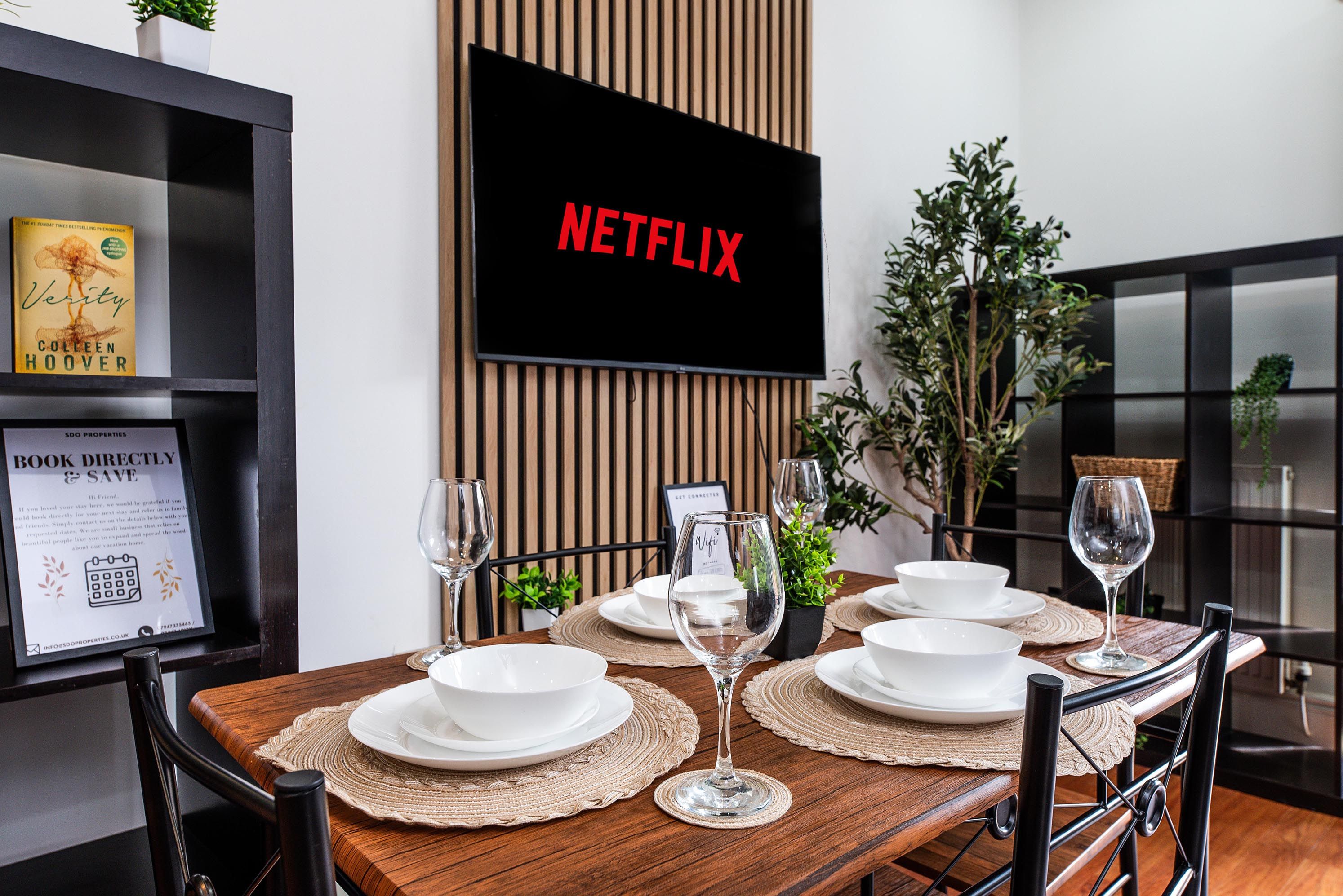 A dining area with a wooden table set for four, a flat-screen TV mounted on a wooden panel, and a framed book on the left.
