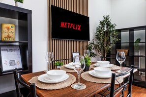 Un comedor con una mesa de madera dispuesta para cuatro, un televisor de pantalla plana montado en un panel de madera y un libro enmarcado a la izquierda.
