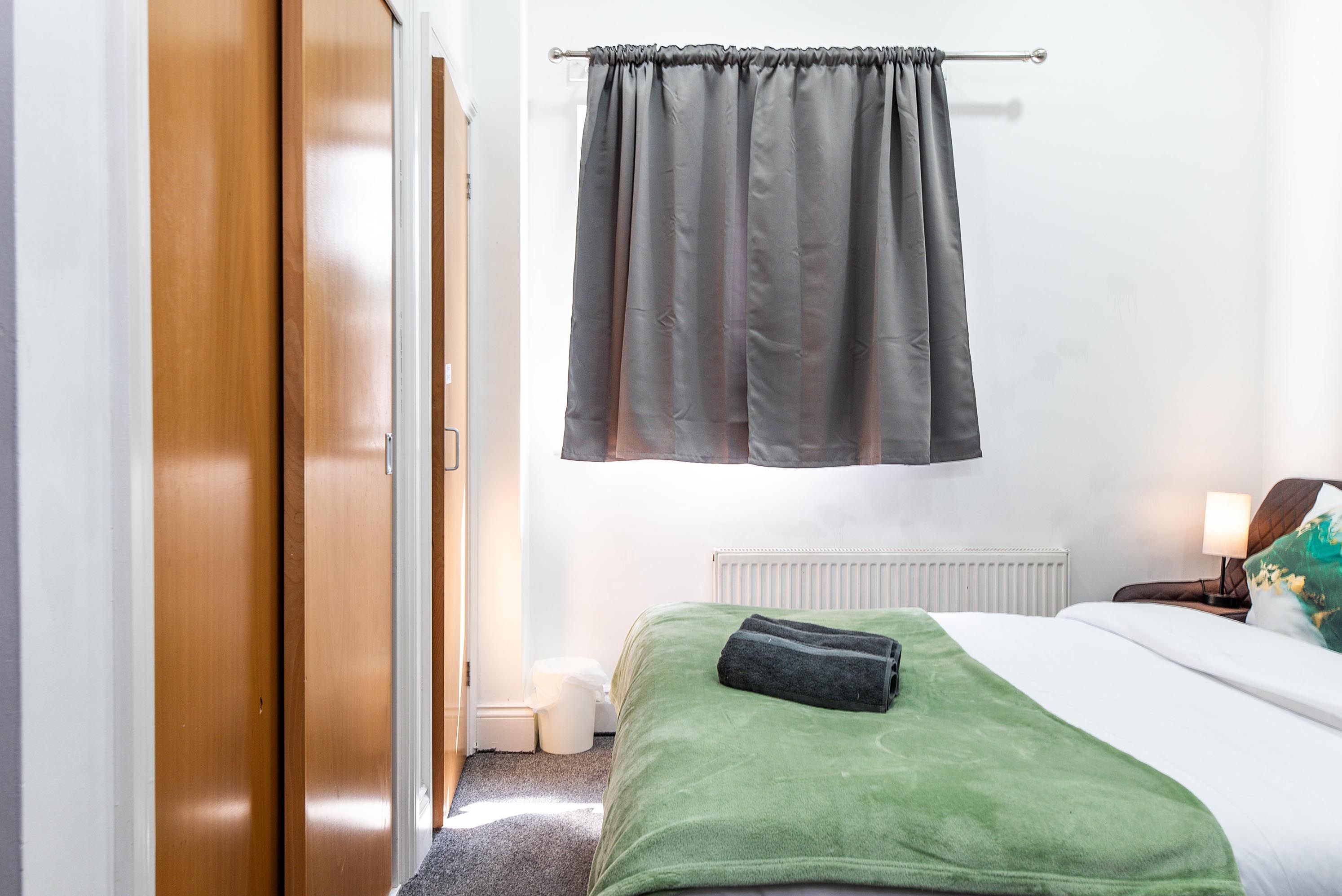 Une chambre d’hôtel avec un lit, une couverture verte, un rideau gris, un radiateur blanc et une porte en bois.