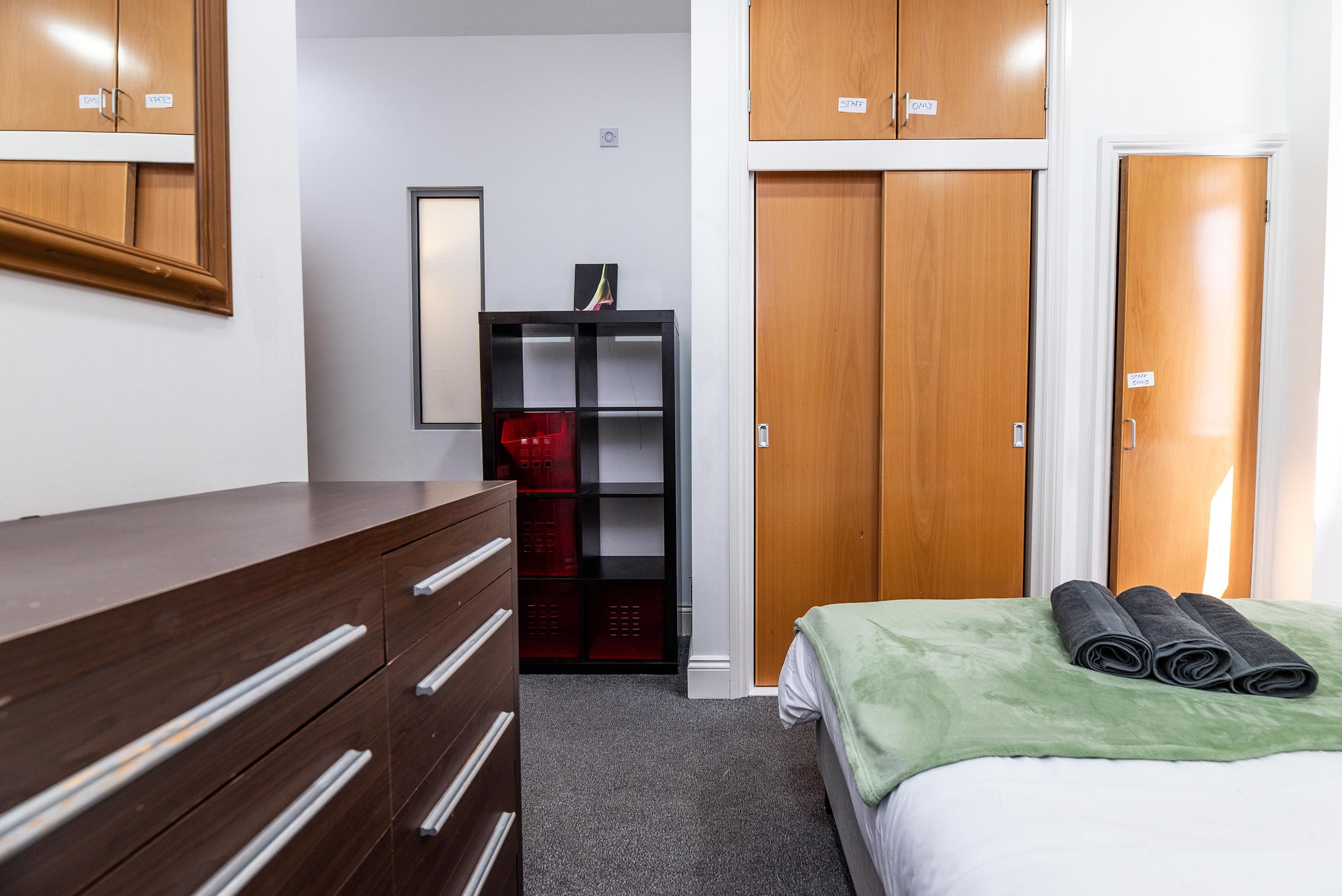 A hotel room with a wooden dresser, a bed with a green blanket, a red storage unit, and a mirror on the wall.