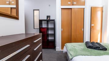 A hotel room with a wooden dresser, a bed with a green blanket, a red storage unit, and a mirror on the wall.