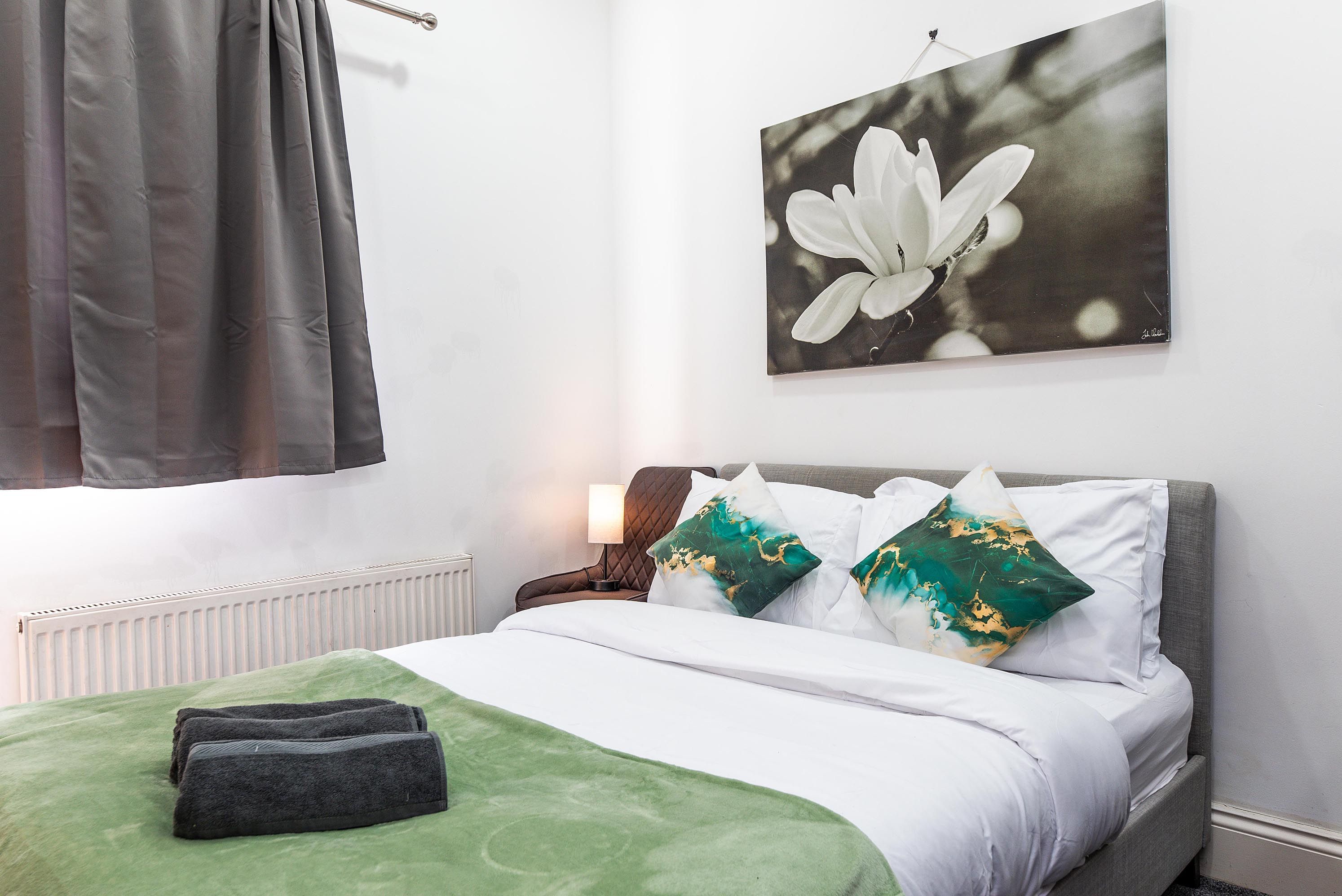 A hotel room with a bed, a radiator, a window with curtains, and a wall-mounted picture of a flower.