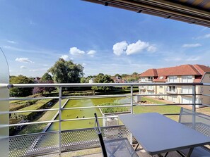A balcony with a view of a pond, a building with a red roof, and a clear blue sky. - Green Lake Retreat with balcony by Interhome (De Haan)