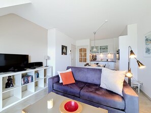 A modern living room with a flat-screen TV, a sofa, a coffee table, and a kitchen in the background. - Green Lake Retreat with balcony by Interhome (De Haan)