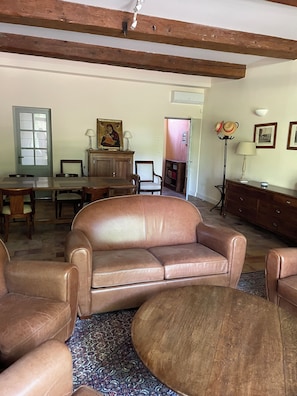 A living room with a brown leather sofa, wooden dining table, and chairs. There is a patterned rug on the floor.