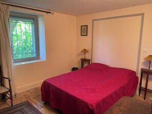 Una habitación con cama, una ventana con cortinas, una mesita con lámpara y un cuadro en la pared.