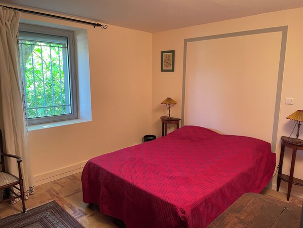A room with a bed, a window with curtains, a small table with a lamp, and a picture on the wall.
