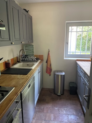 Una cocina con armarios grises, un lavamanos de acero inoxidable, una ventana con vistas a zonas verdes y un cubo de basura.