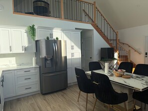 Une cuisine moderne avec des meubles blancs, un réfrigérateur en acier inoxydable, une table à manger avec des chaises noires et un escalier en bois.