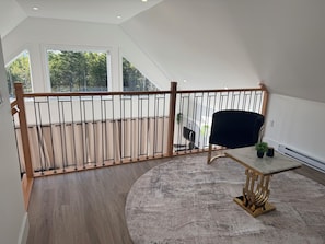 A spacious room with a high ceiling, a wooden railing, a round rug, a table with a gold base, and a black chair.