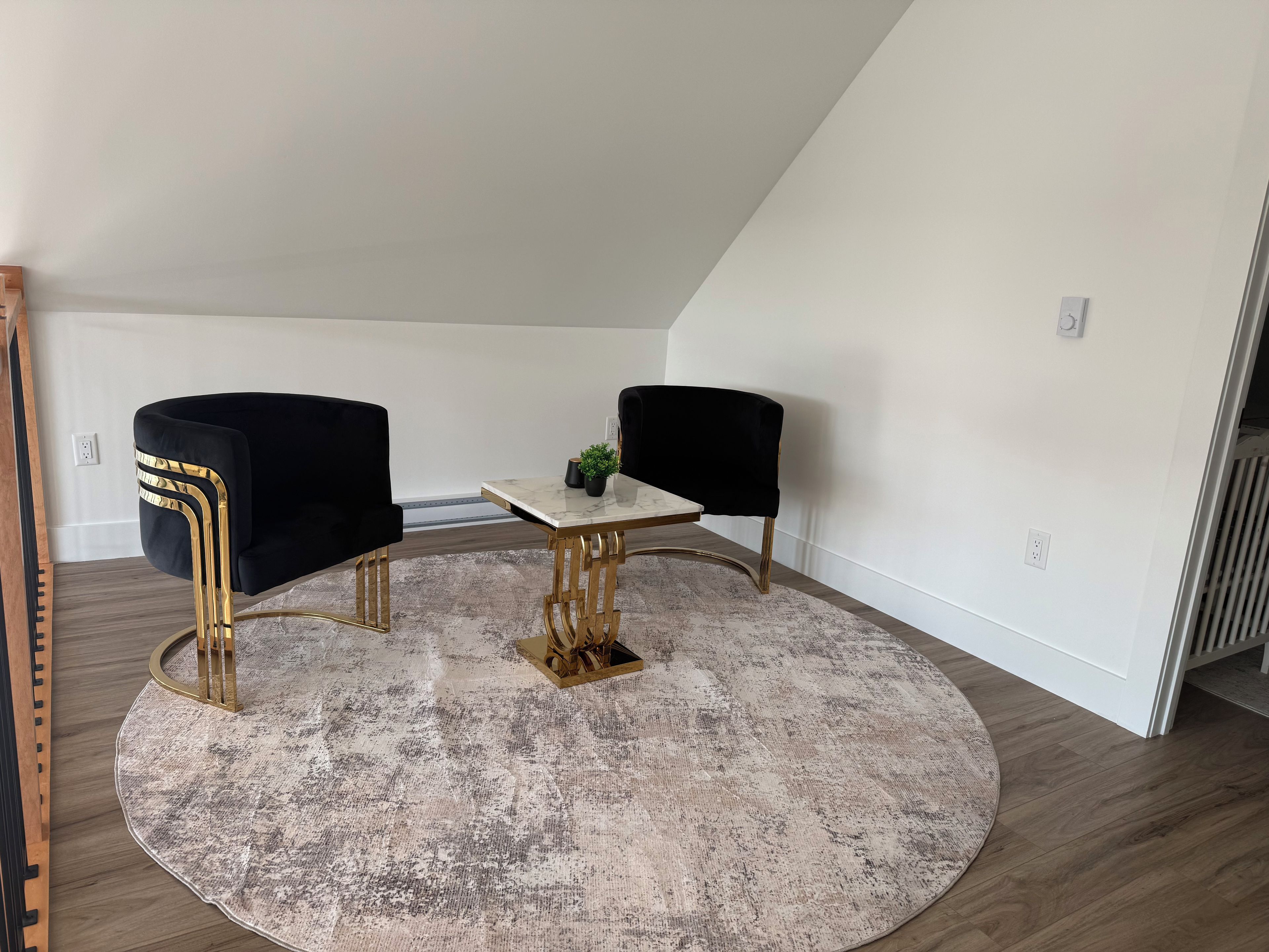 A room with a round, distressed rug, a small table with a marble top, and two black chairs with gold legs.