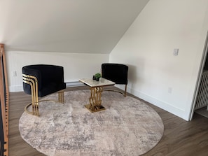 A room with a round, distressed rug, a small table with a marble top, and two black chairs with gold legs.