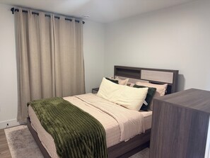 Un lit bien fait, avec une couverture verte, une tête de lit en bois et une table de chevet surmontée d’une lampe.