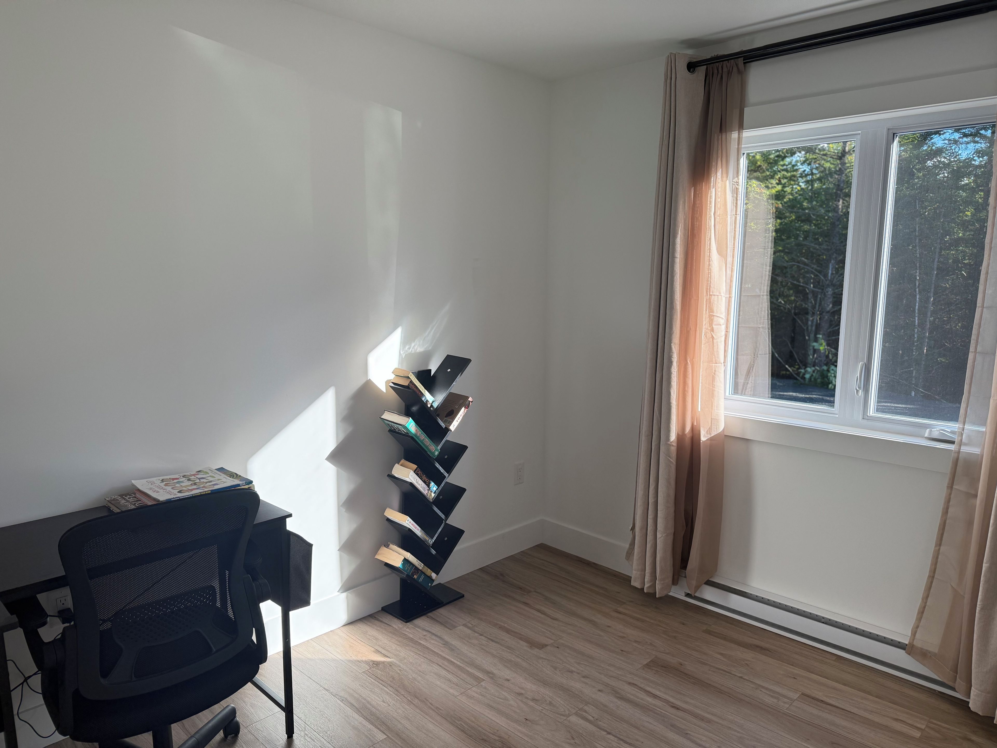 A room with a black office chair, a wall-mounted bookcase, a window with curtains, and a wooden floor.